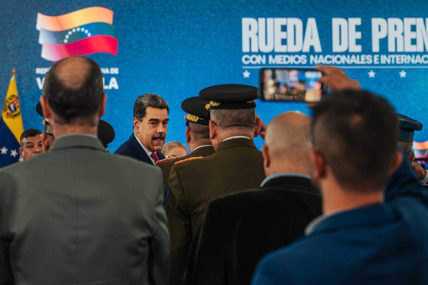 Nicolás Maduro facing the camera while being saluted and photographed by a group of men, some in military uniform.