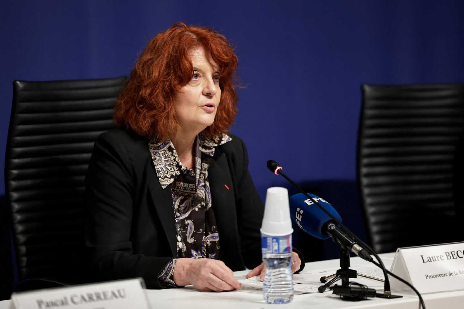 A woman with reddish hair sits at a table before a microphone and a water bottle. 
