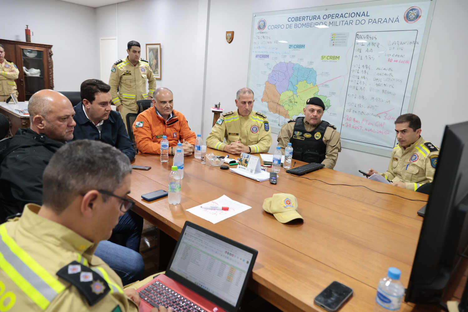 A group of men sitting around a conference table, some wearing emergency worker jackets, one holding a laptop.