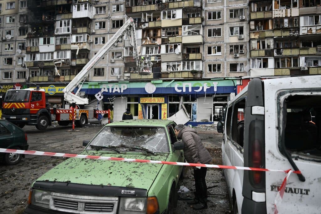A person leans into a green car with a shattered windshield, behind red and white caution tape. A red fire truck extends its ladder toward a multistory building with broken windows.