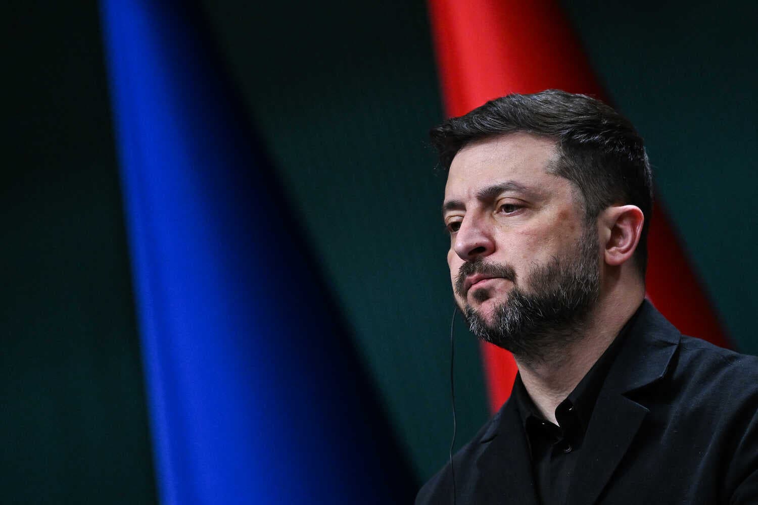 Volodymyr Zelensky, with dark hair and a beard, looks ahead. He wears a dark jacket against a dark background with blue and red bands.