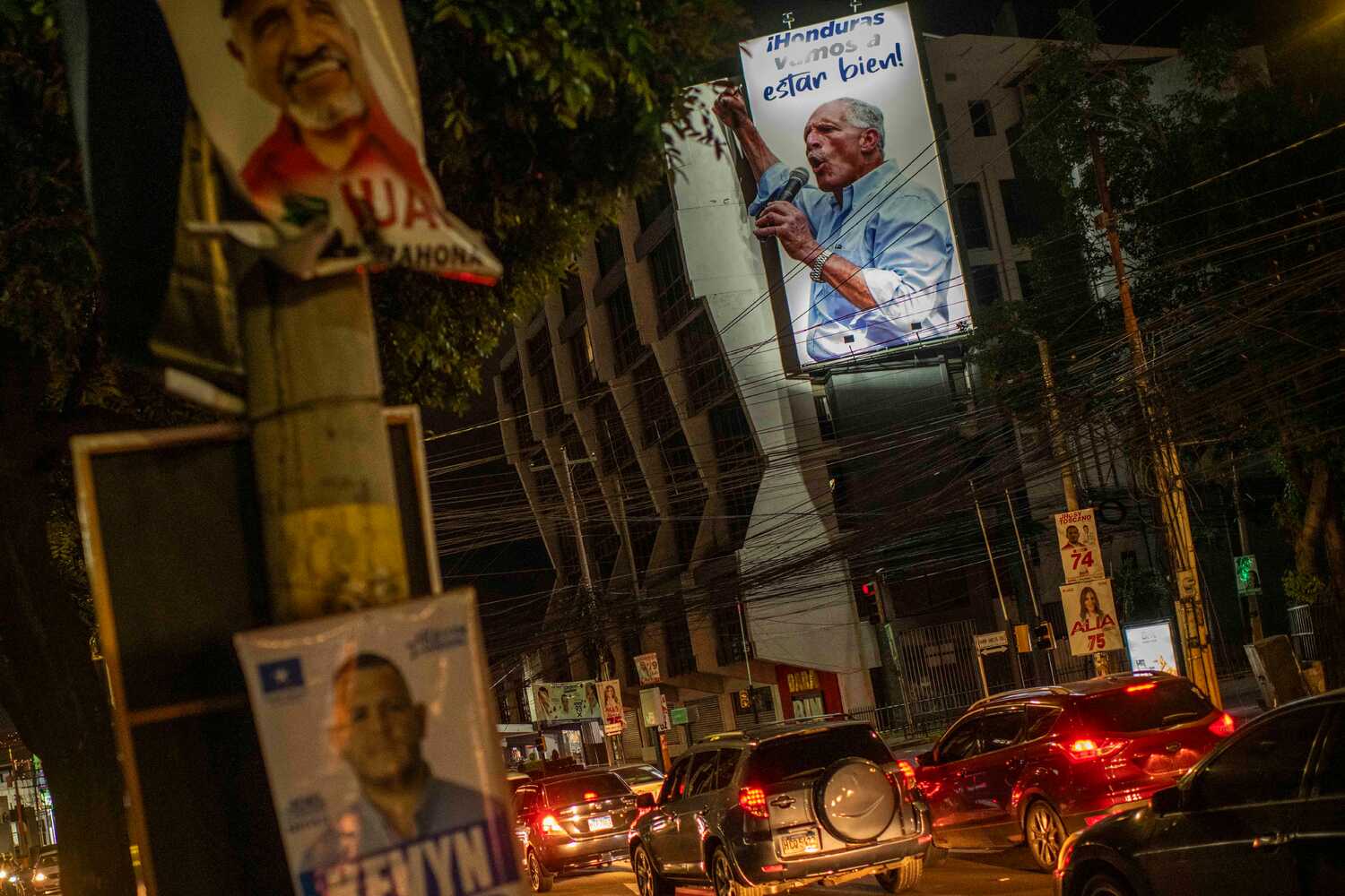 A poster of a man in a blue shirt speaking into a microphone hangs above traffic on a city street.