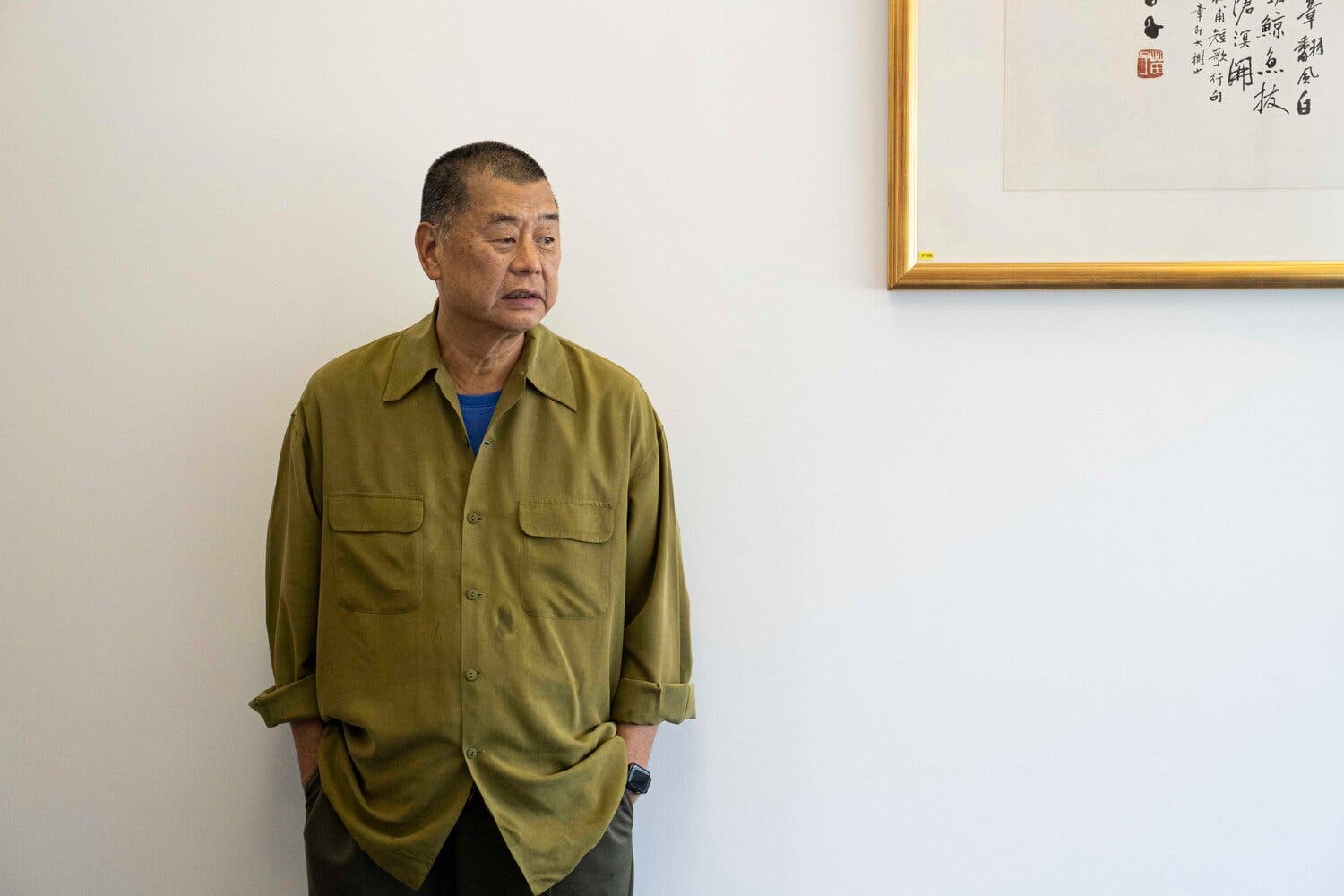 Jimmy Lai stands in an olive shirt with his hands in his trouser pockets. Behind him is a white wall and the corner of a framed picture.