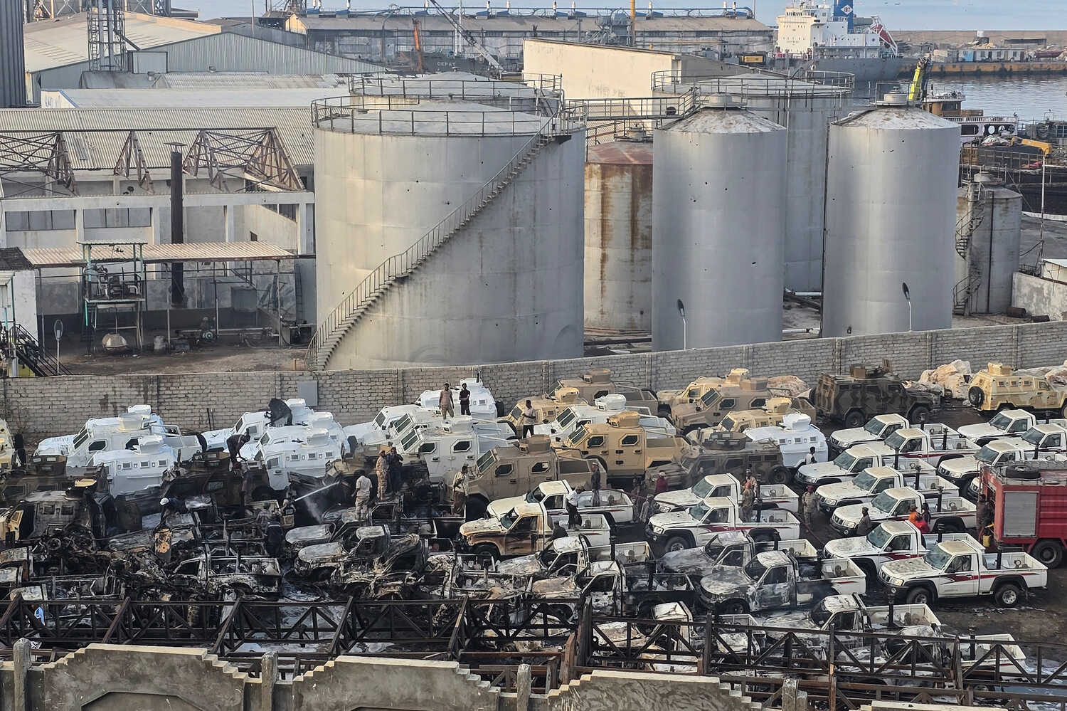 A vast industrial yard is filled with charred remnants of vehicles and some intact trucks. A few people are standing among the vehicles, with large industrial tanks and a body of water visible in the distance.