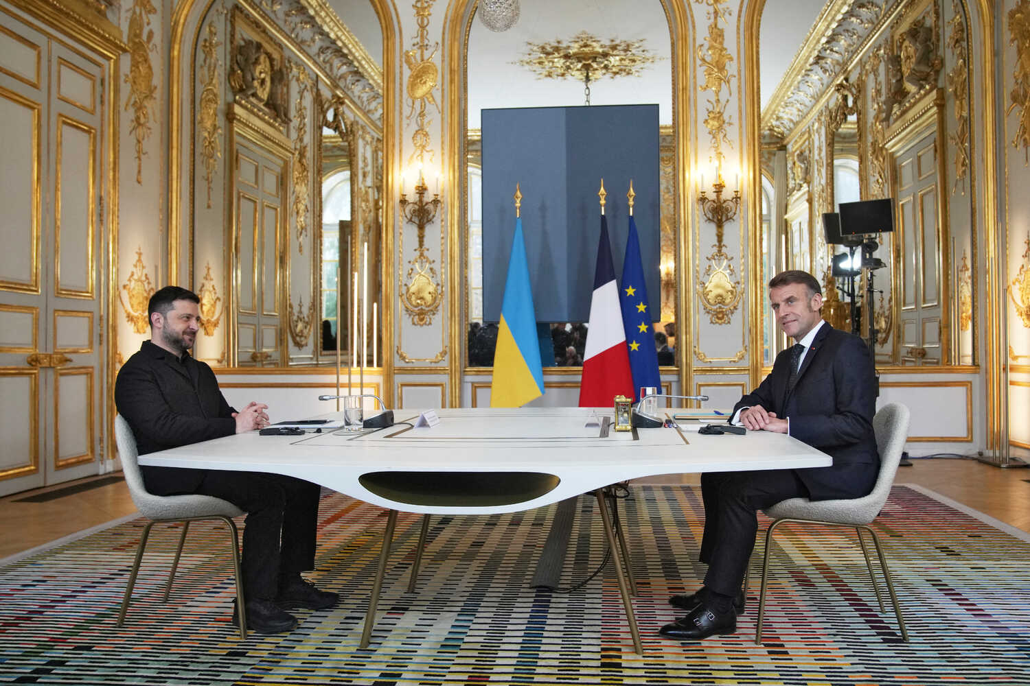 Volodymyr Zelensky and Emmanuel Macron of France sit at a table together, with flags in the background.
