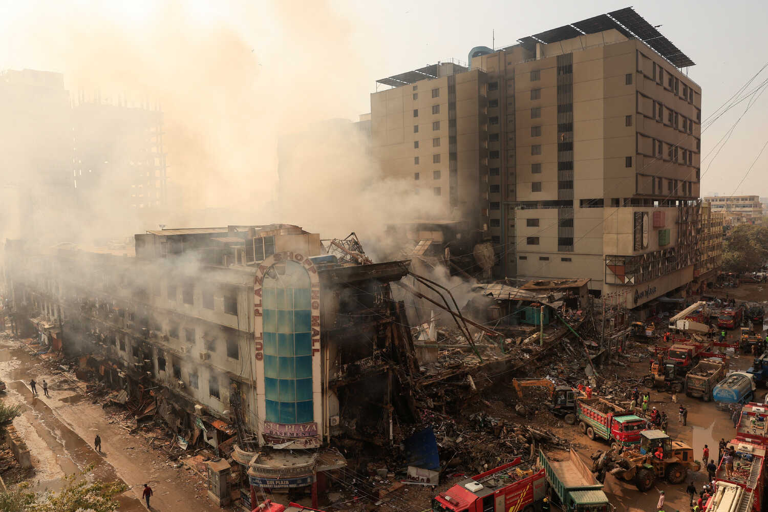 Clouds of smoke rise above a partially collapsed multistory building. Emergency vehicles can be seen next to the rubble. 