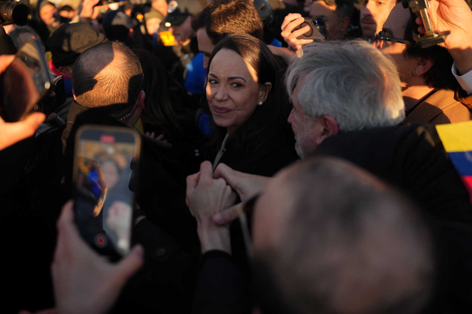 María Corina Machado, wearing a dark coat and pearl earrings, smiles while turning her head in a crowded setting as people around her hold up phones and cameras.