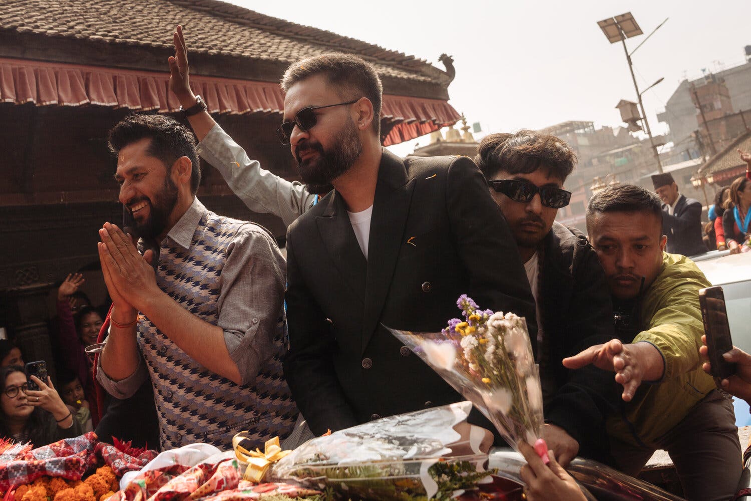 A person in a black suit, sunglasses, and a beard. Bouquets of flowers are next to him.