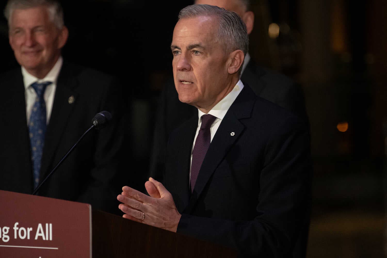 Mr. Carney, wearing in a dark suit, standing at a microphone. Another person in a dark suit smiles in the background.
