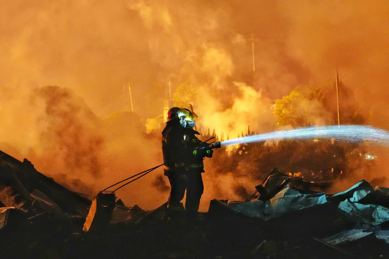 Two people in dark firefighter gear, standing on top of dark debris, spray a stream of water from a hose. Dense orange and yellow smoke fills the frame.