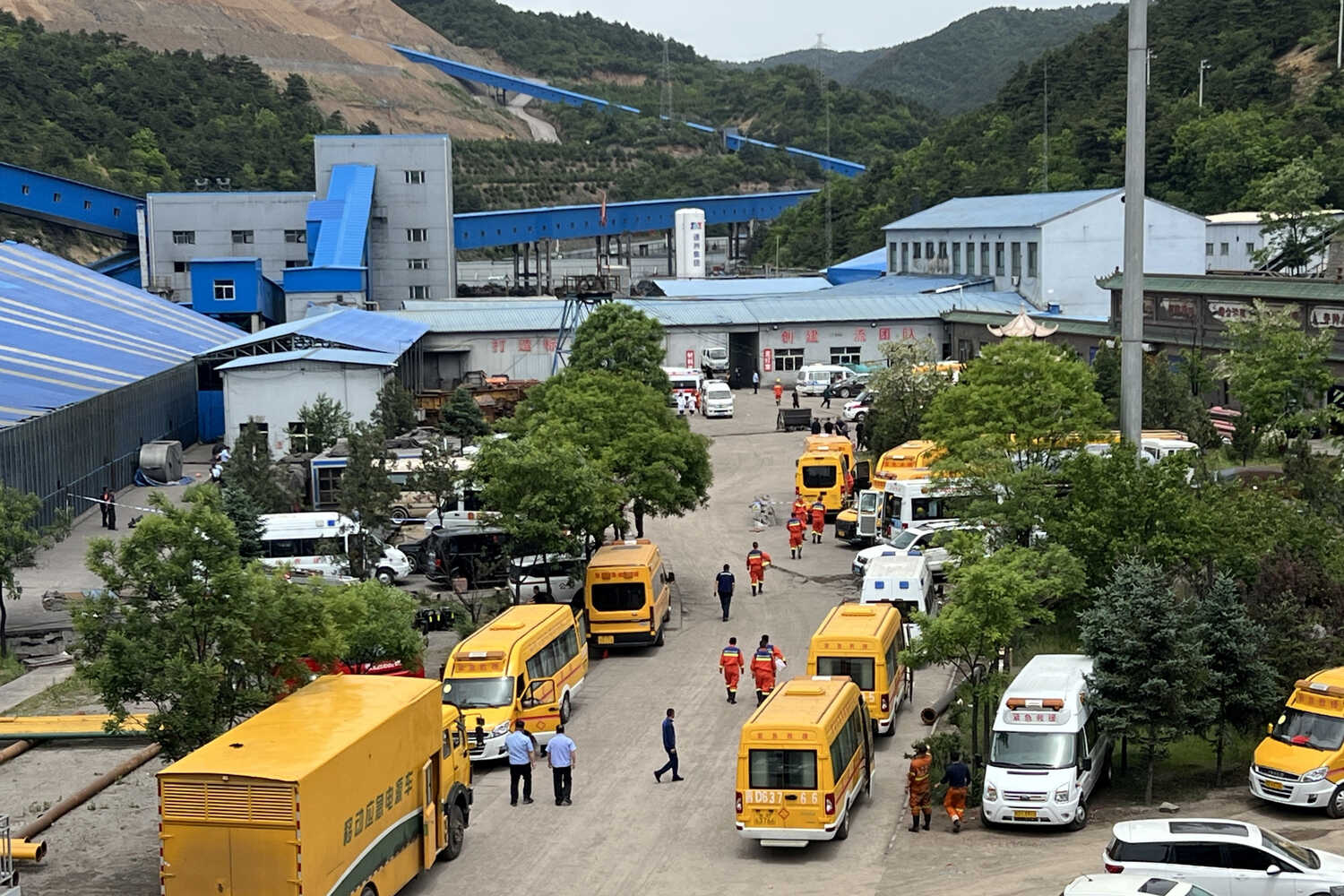 An industrial complex at a mountain’s base, with blue buildings. Many yellow buses, white vans and people in orange uniforms are in the foreground.