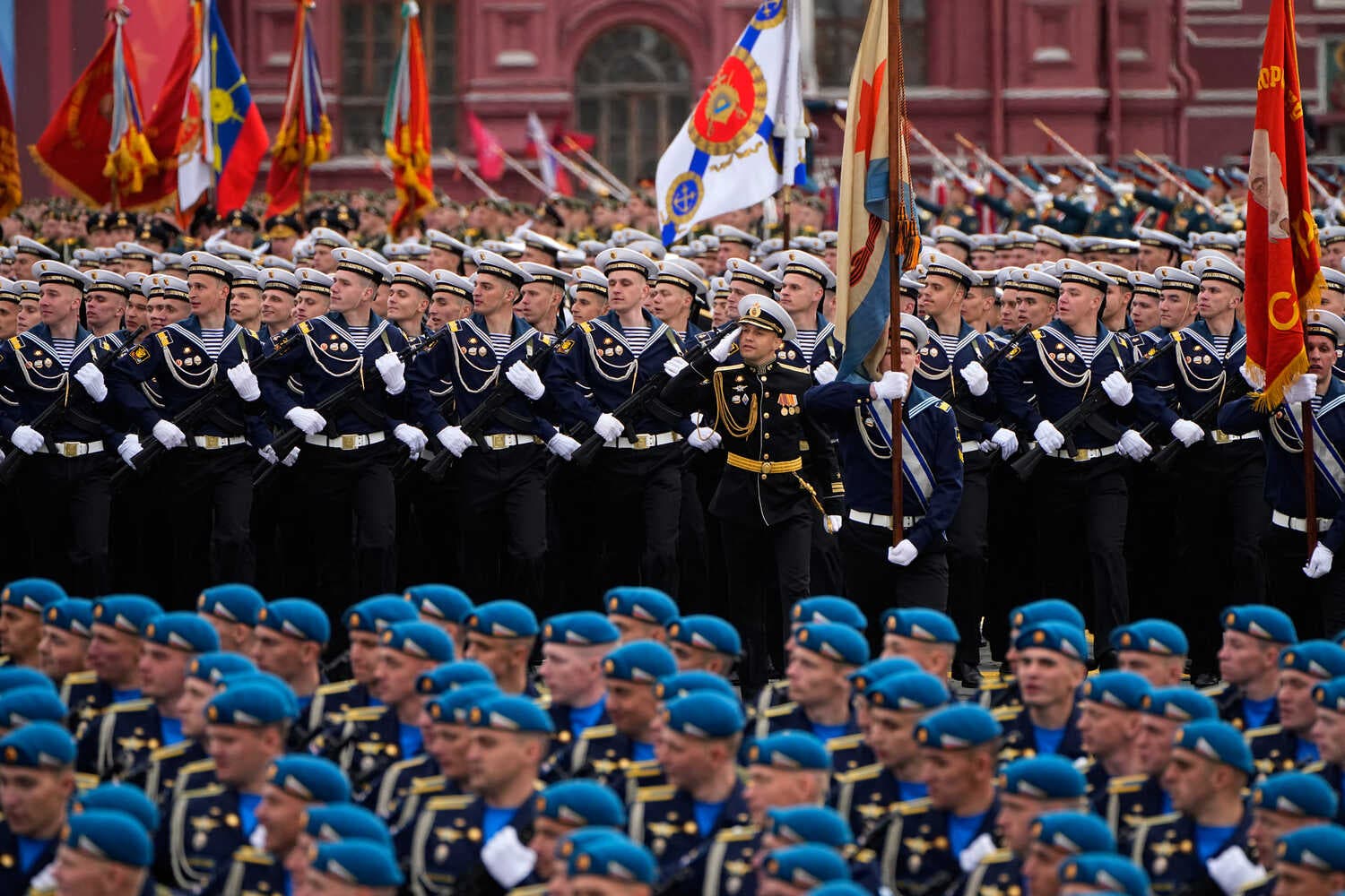 Many people in blue berets and blue uniforms. Behind them, people in dark blue sailor uniforms march in formation, holding guns and flags.