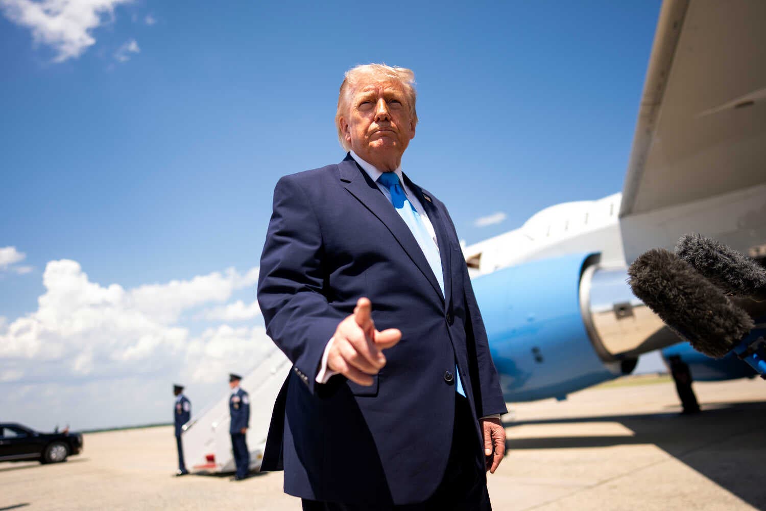 Trump in a navy blue suit and light blue tie points while standing near a plane.