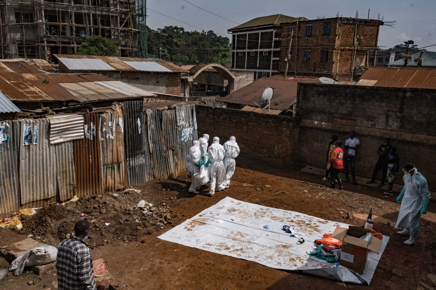 An open dirt area with buildings and corrugated metal structures. Several people in white protective suits are gathered, while another person in a light blue suit and apron stands near a white tarp and box.