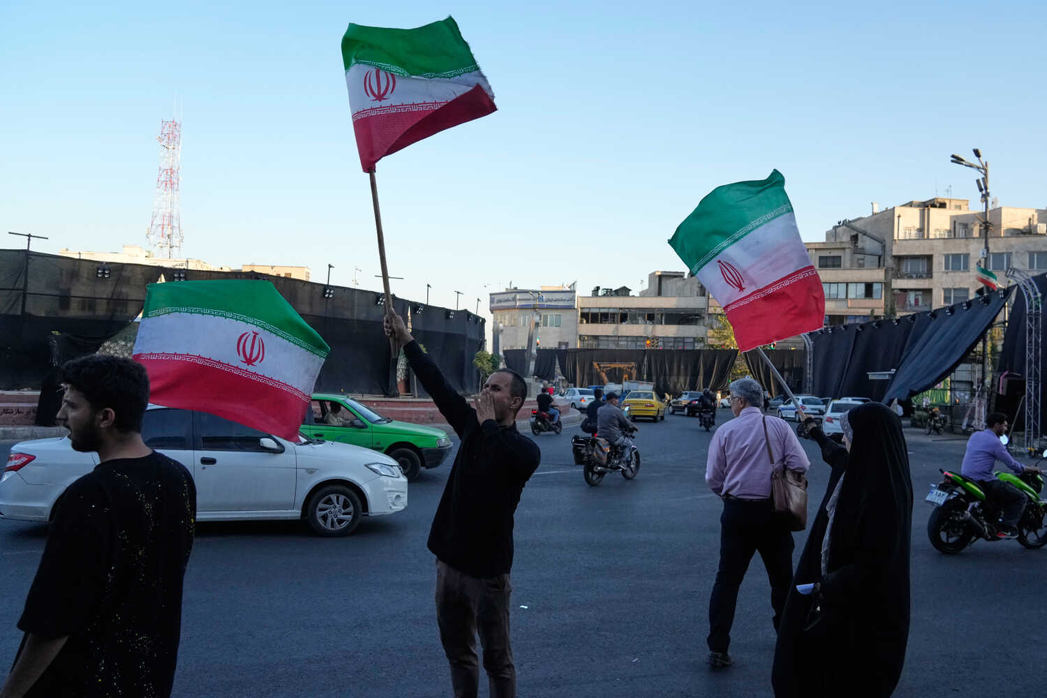People on a street waving red, white and green flags as cars pass by.