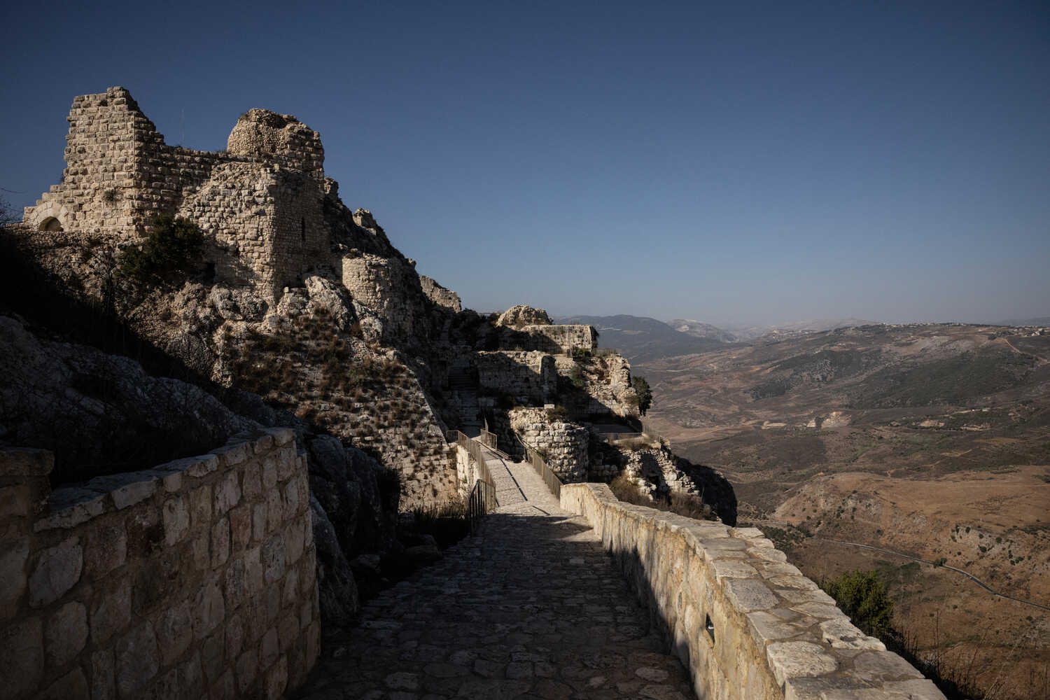 Ancient stone ruins on a high, rocky slope. A winding path and wall lead toward them, overlooking a sweeping landscape of dry hills.